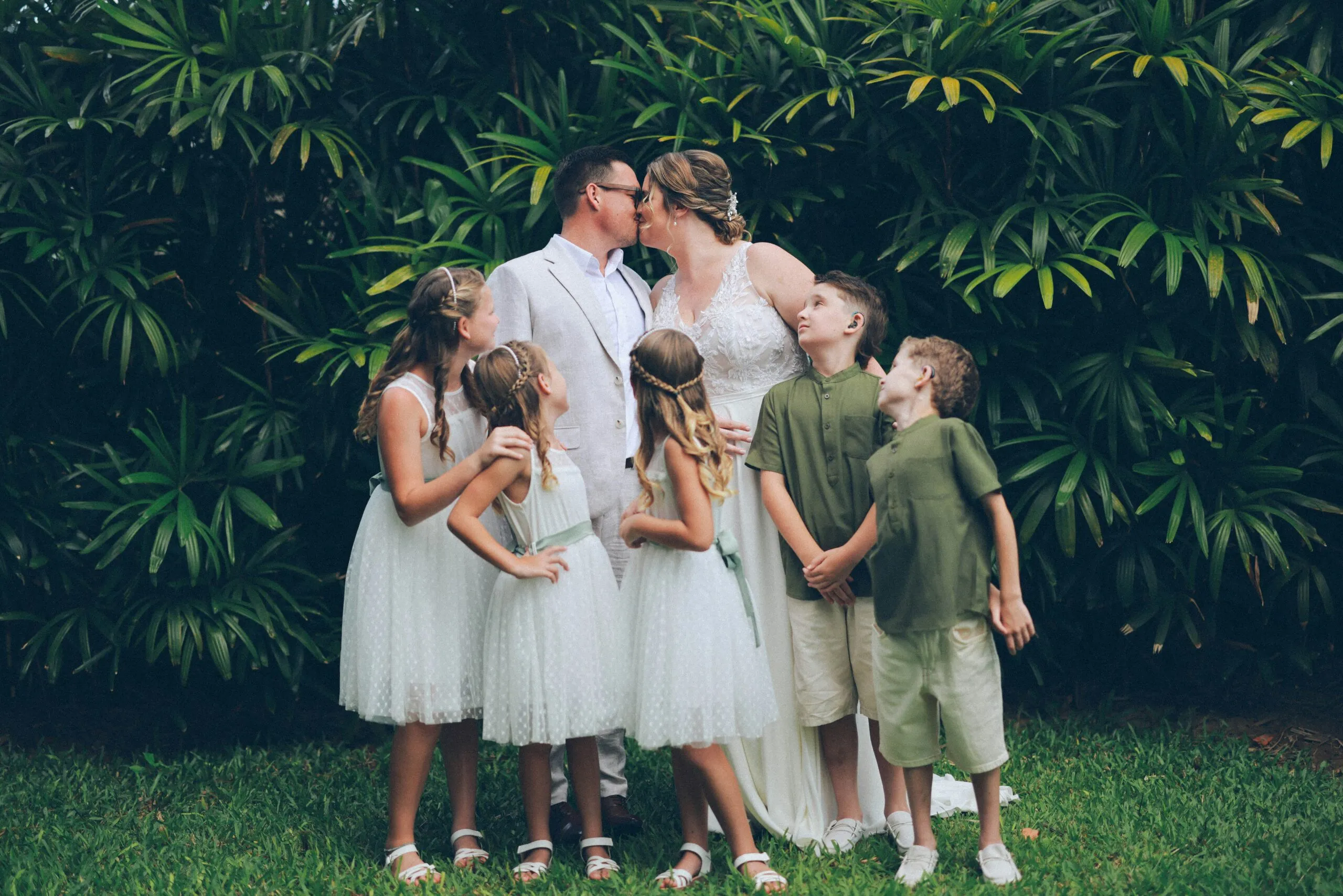 Newlyweds kiss surrounded by children in garden.