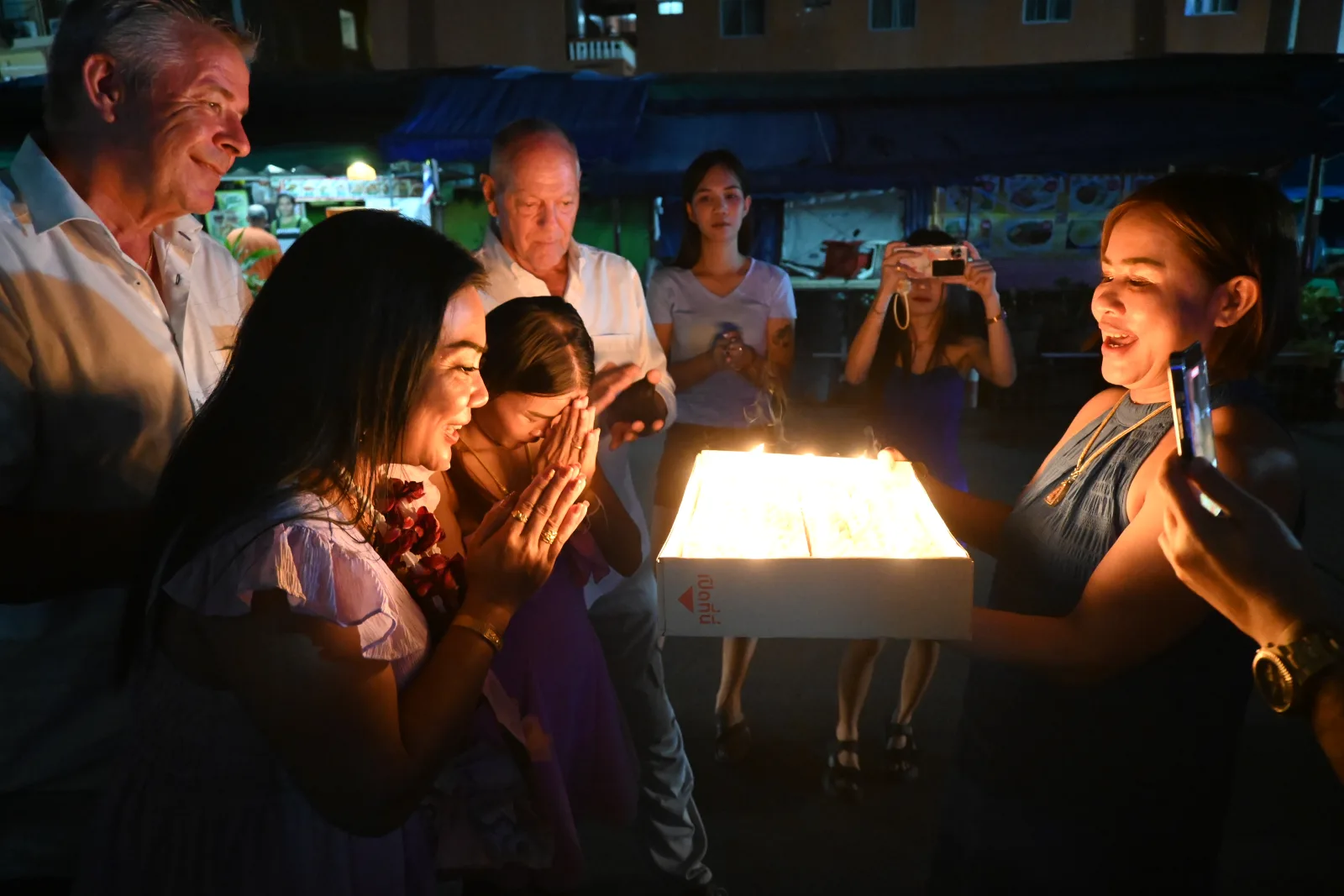The photo shows a nighttime gathering where several people are gathered around an illuminated object, possibly a cake with lit candles. The warm glow from the light source casts a gentle illumination on the faces of those standing around it. Some individuals have their hands pressed together in what appears to be a gesture of prayer or celebration, while others are watching the scene. One person in the background seems to be capturing the moment with a camera or phone. The setting looks like it might be outdoors with buildings visible in the dimly lit background. This appears to be a celebration or ceremonial moment, perhaps a birthday or cultural event, with the light creating a warm, intimate atmosphere in the darkness.