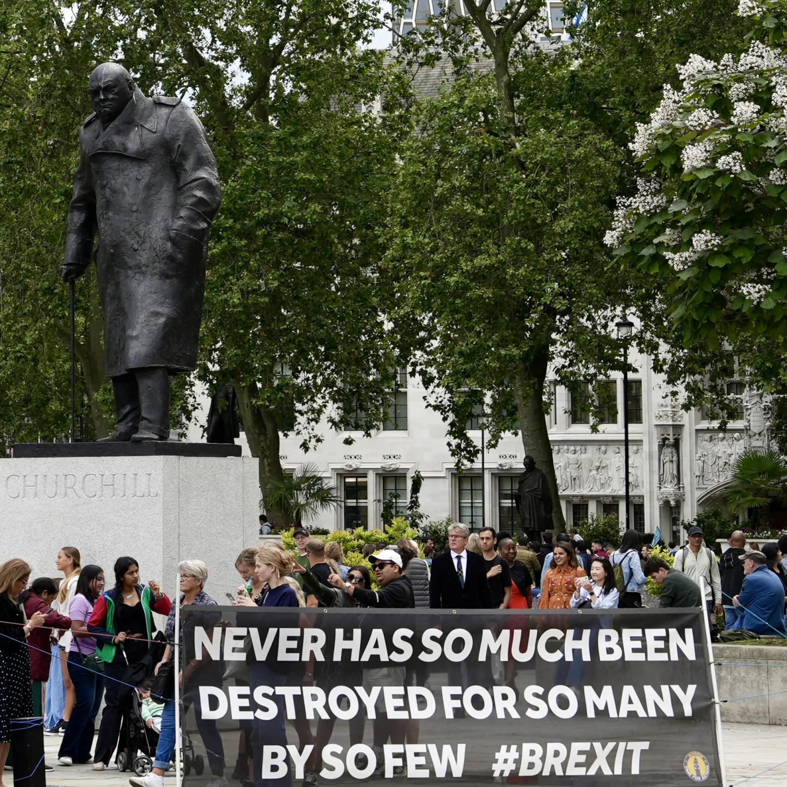 A diverse crowd walks between a protest banner and the statue of Winston Churchill before the Supreme Court of the UK.
