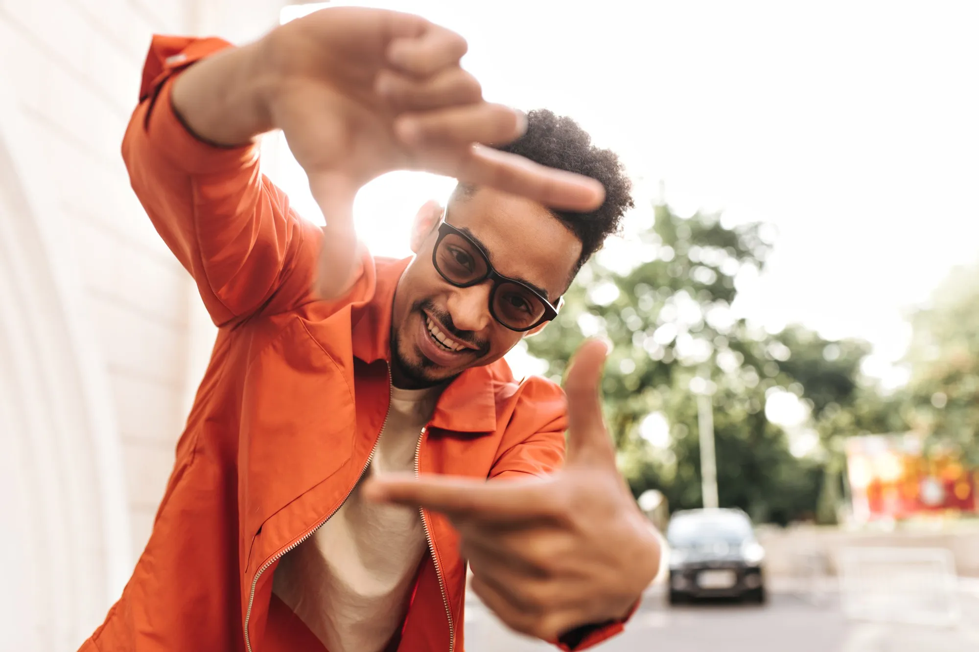 Cool excited curly dark-skinned man in bright orange jacket and sunglasses smiles and makes camera sign with his fingers outside