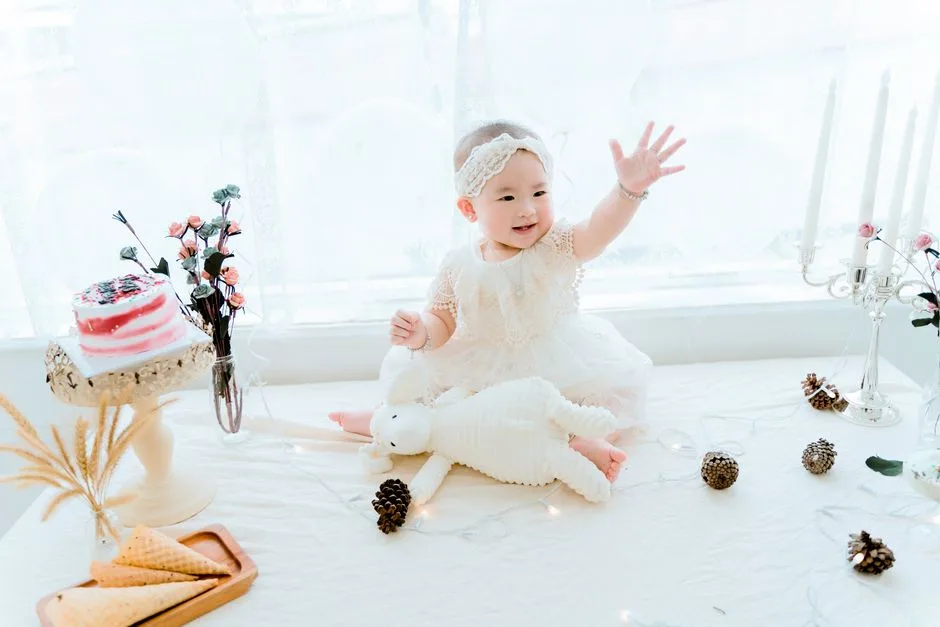 Cute baby girl in white dress celebrating her birthday indoors with cake and toys.