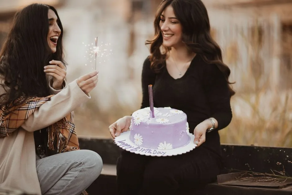 Two women celebrating a birthday outdoors, smiling with a purple cake and sparklers.