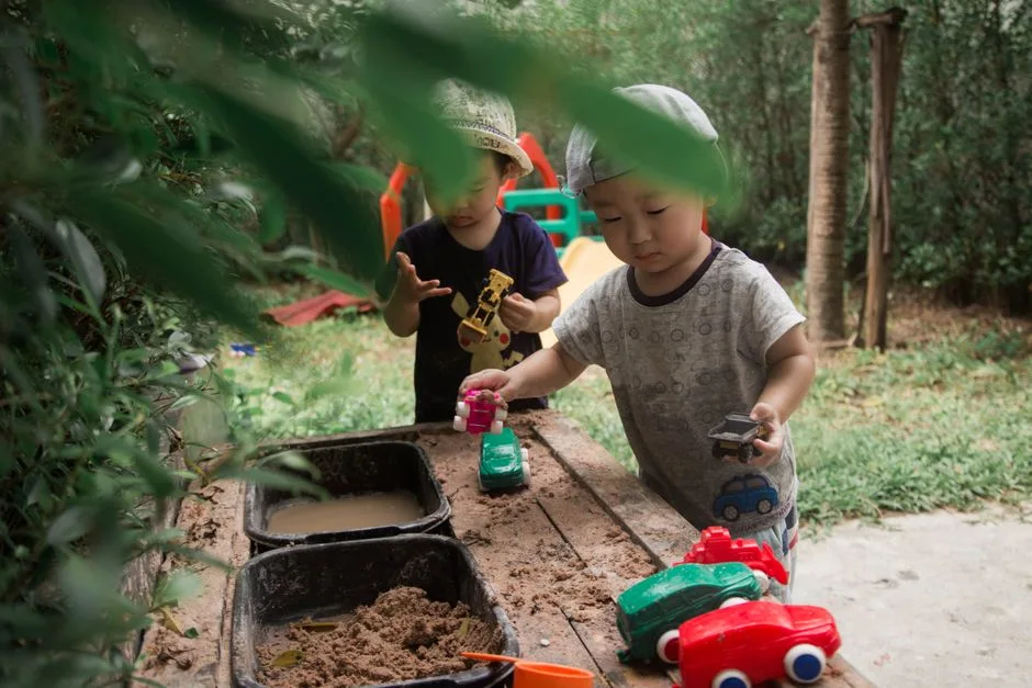 Young children engaged in imaginative play with toy cars in a garden sandbox.