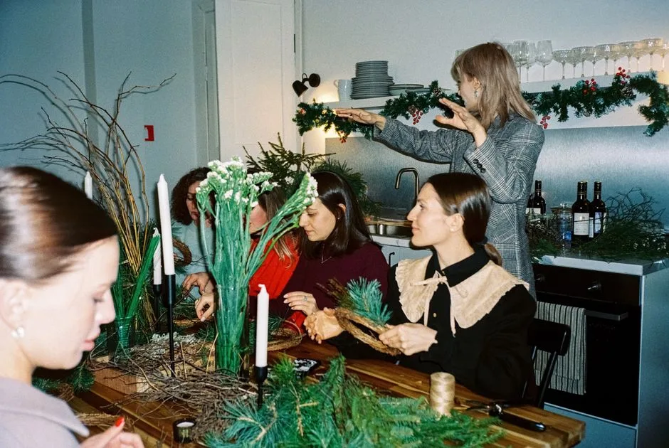 Women crafting festive wreaths at a cozy indoor workshop, surrounded by evergreen branches and candles.