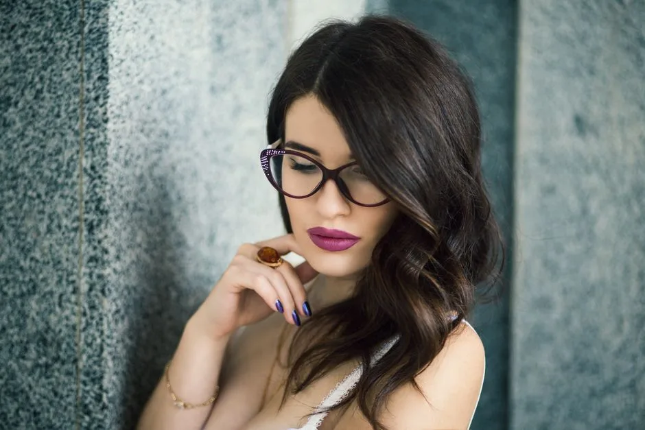 Portrait of an elegant woman with glasses and bold lipstick, posing against a stone background.