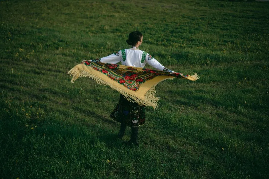 A woman wearing traditional attire with a colorful shawl stands on a grassy field, capturing cultural essence.