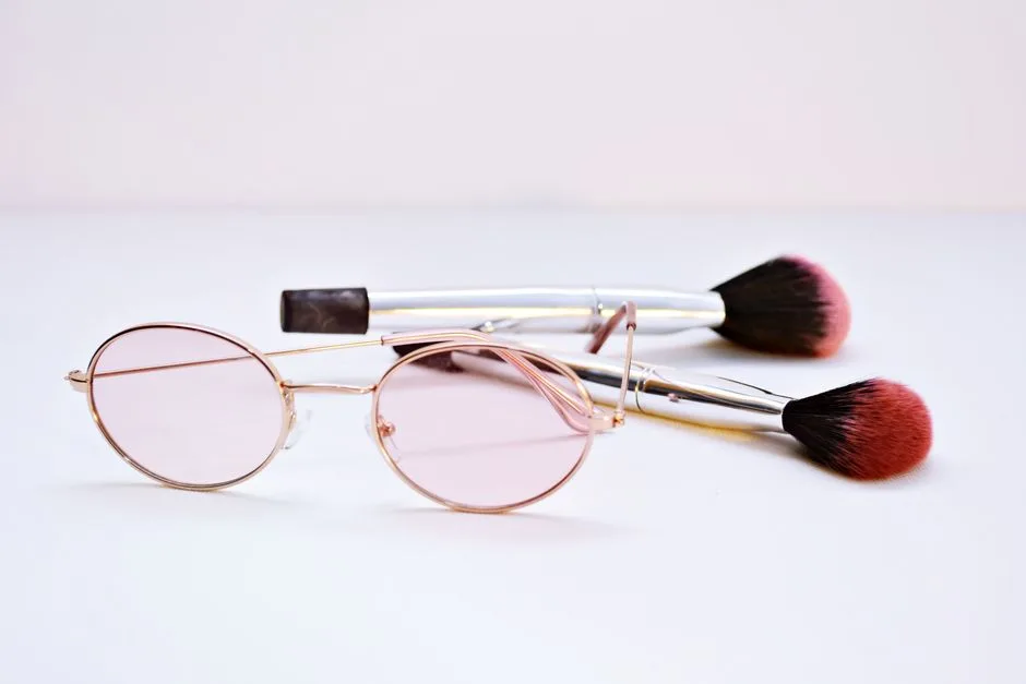 Stylish eyeglasses with pink lenses and makeup brushes on a neutral background.