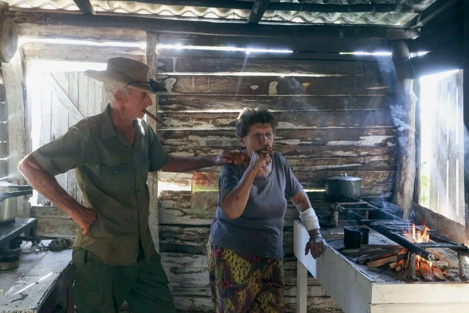 Elderly couple enjoying cigars in a rustic wooden shed. Smoky ambiance, natural light filtering in.