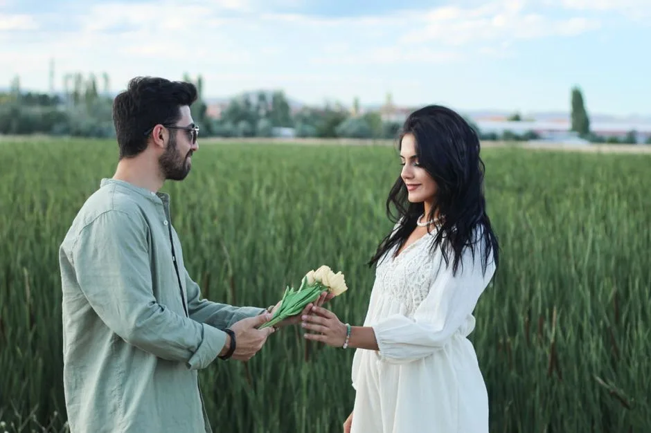 A man and woman share a bouquet, expressing love in a lush green field.
