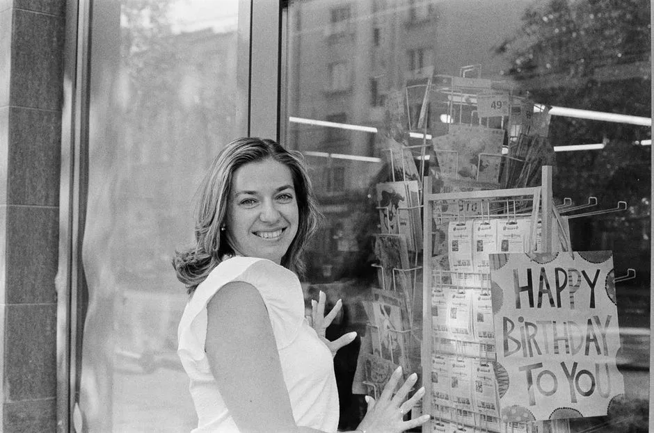 A black and white portrait of a woman smiling near a birthday card display in an urban setting.