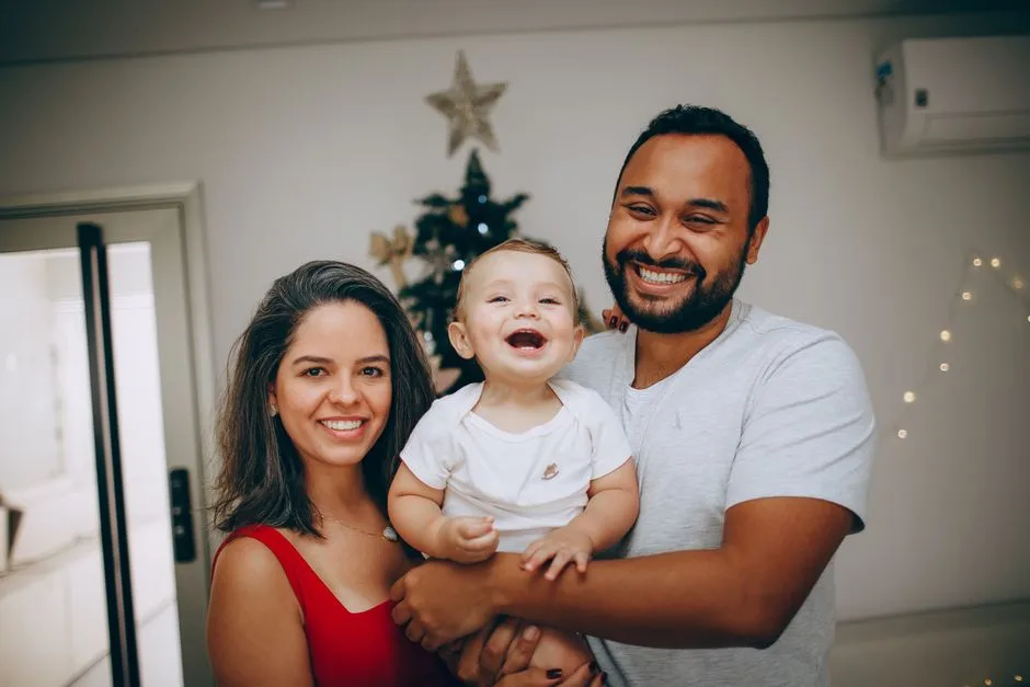 A joyful family of three smiling in front of a decorated Christmas tree indoors.