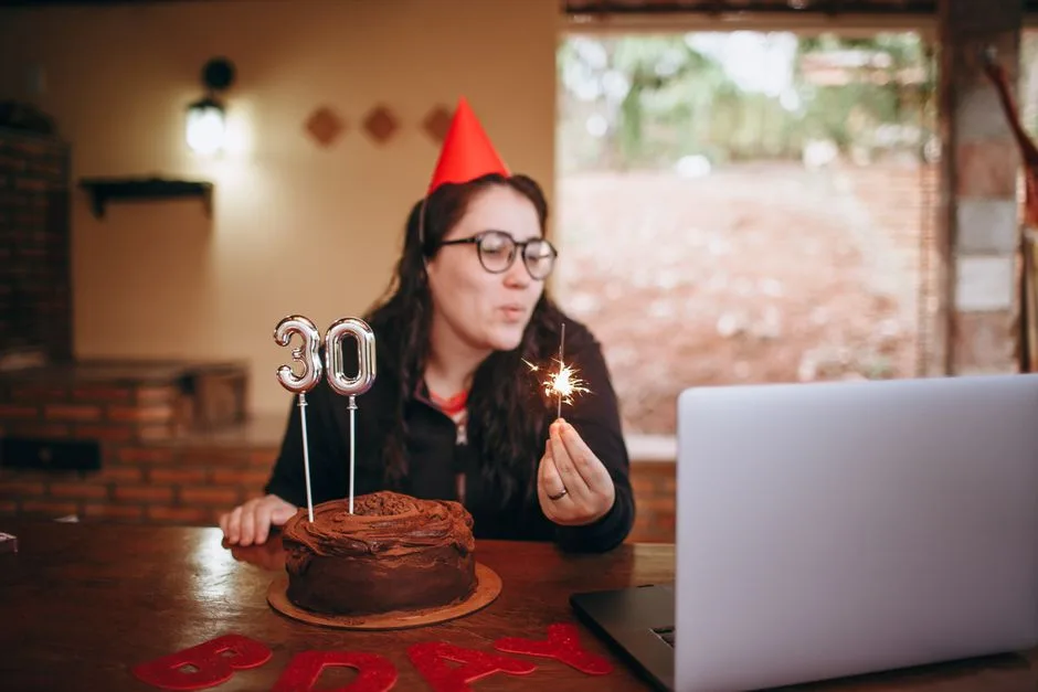 A woman in glasses celebrates her 30th birthday online, blowing out a cake candle, while sitting in a cozy room.