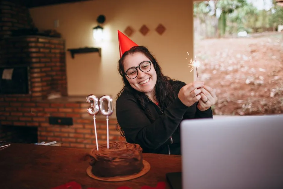 Cheerful woman in party hat holding sparkler with chocolate cake for 30th birthday celebration indoors.