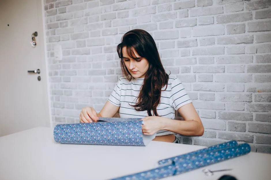 A woman focused on wrapping a gift with floral blue paper at a table indoor setting.