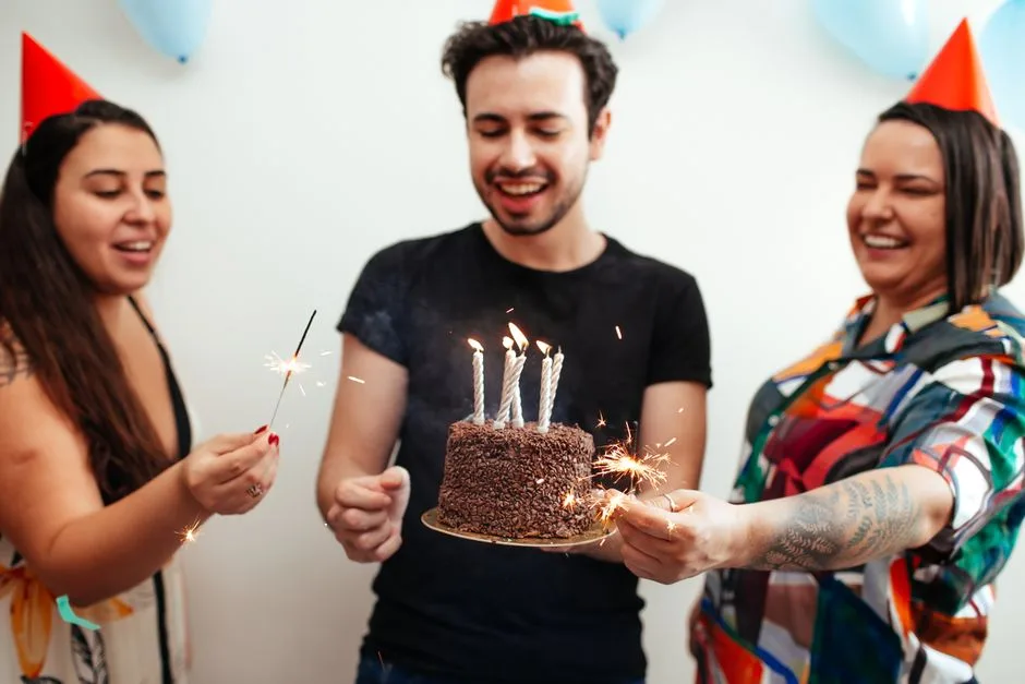 Three friends celebrate a birthday with cake and sparklers indoors wearing party hats.
