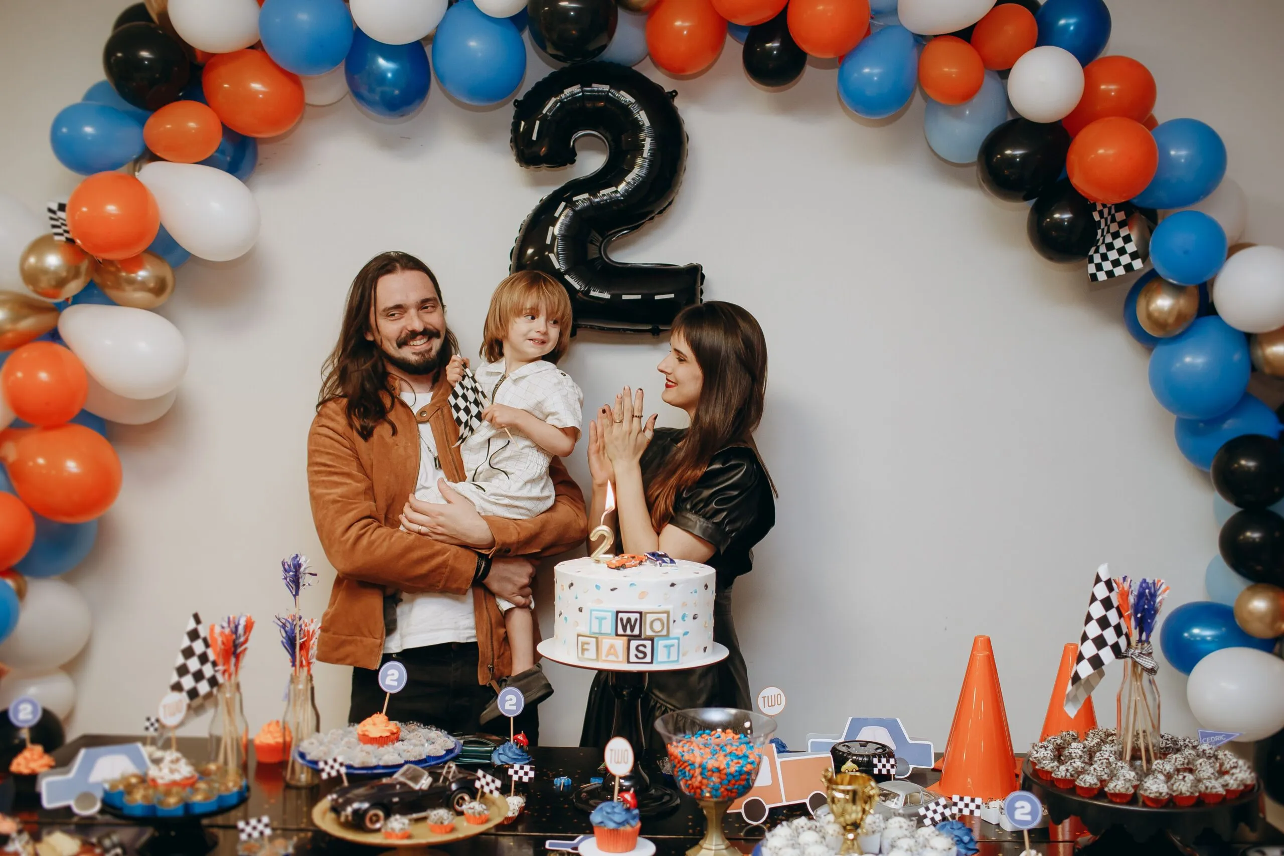A family celebrates a child's second birthday with balloons and cake indoors.