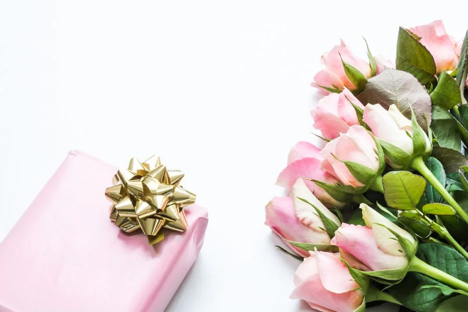A bouquet of pink roses next to a pink gift box with a gold bow on a white background.