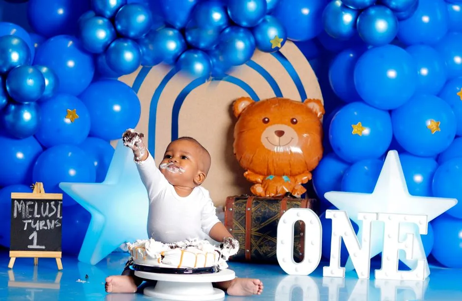 Adorable baby enjoying first birthday cake amidst blue balloon decorations.