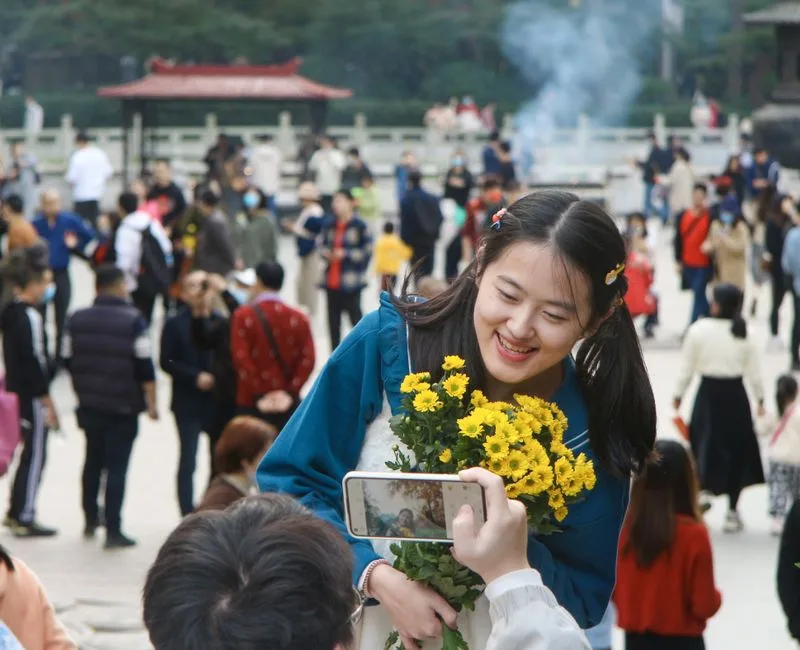 A young woman holding yellow flowers, smiling during an outdoor festival with many people around.