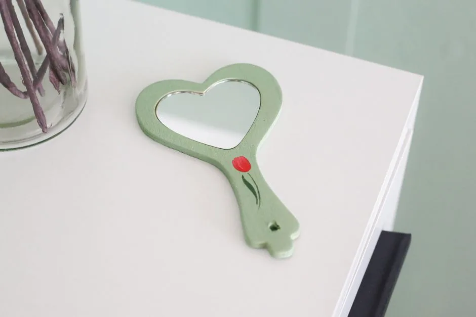 A heart-shaped mirror with a painted red tulip on a minimalist white table.