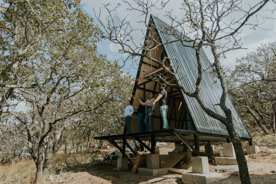 Group constructing modern A-frame cabin in forest surrounded by oak trees.