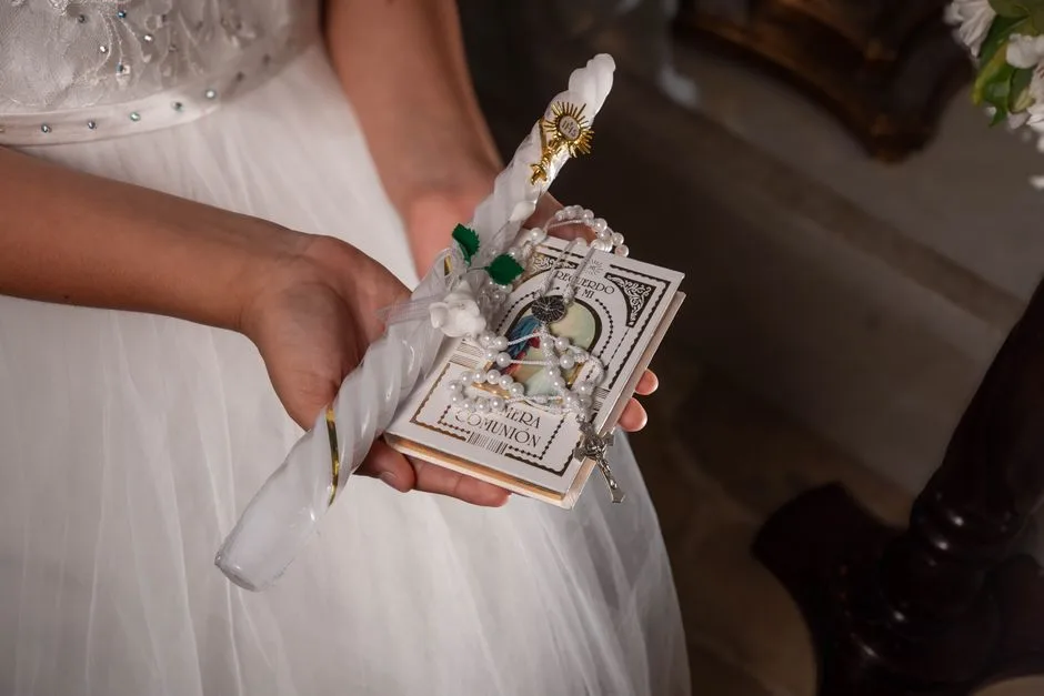 Close-up of hands holding First Communion items including candle, rosary, and book.