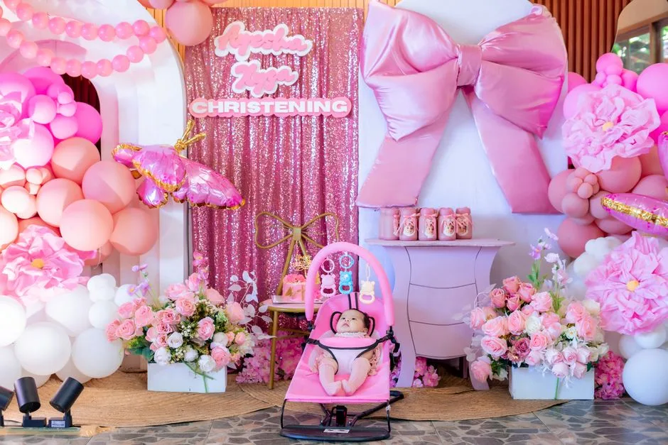 A beautiful pink-themed christening setup with a baby in a seat, floral arrangements, and balloons.
