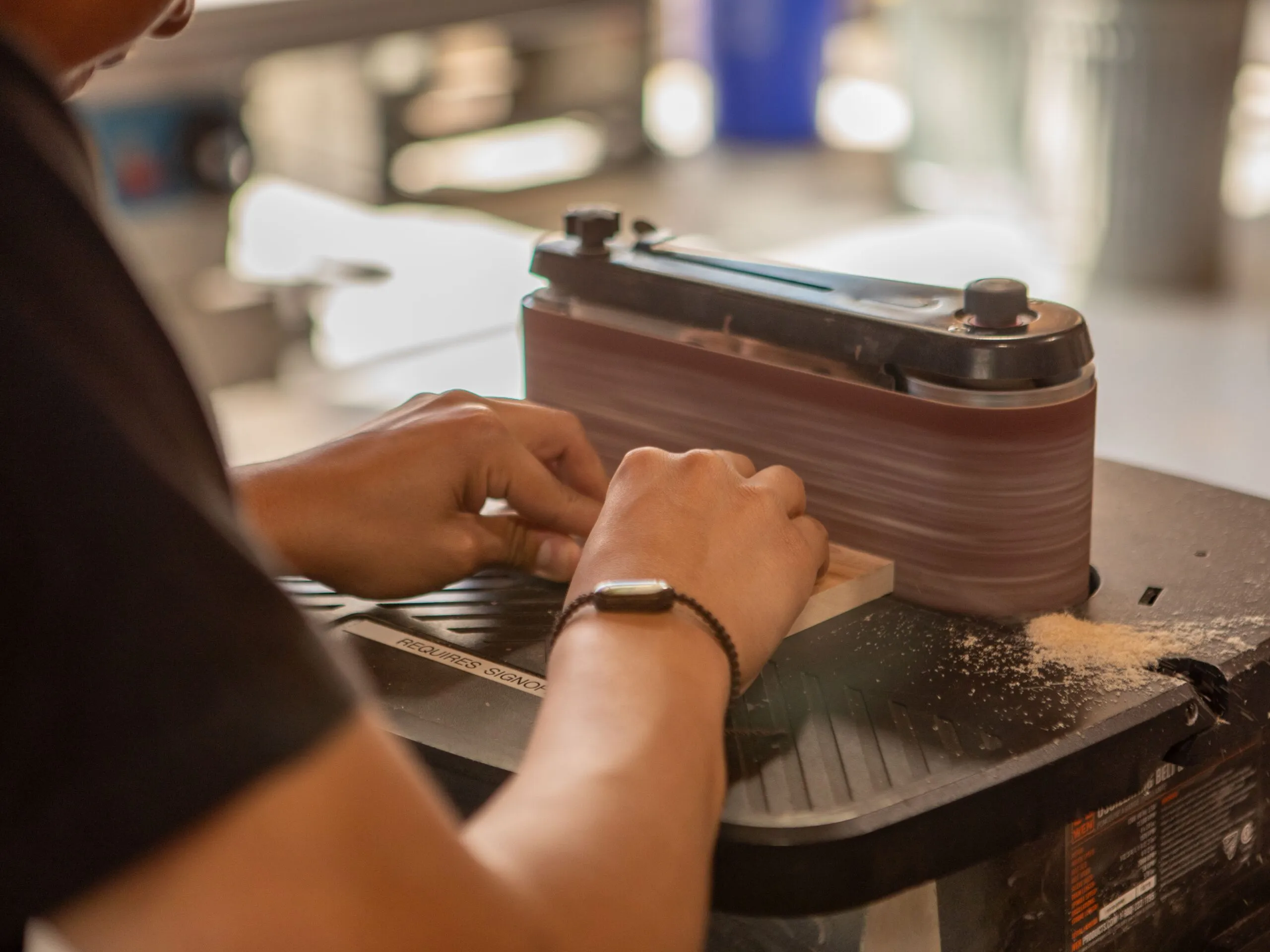 Hands of a craftsman using a belt sander to smooth wood in a workshop setting.