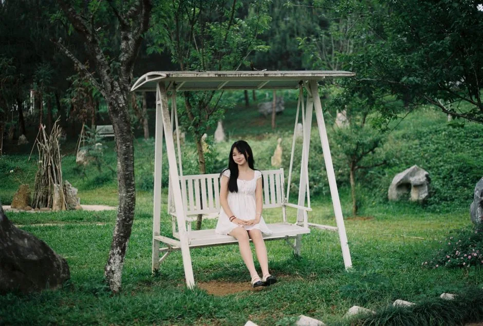 A young woman in a white dress sits on a garden swing surrounded by lush greenery.
