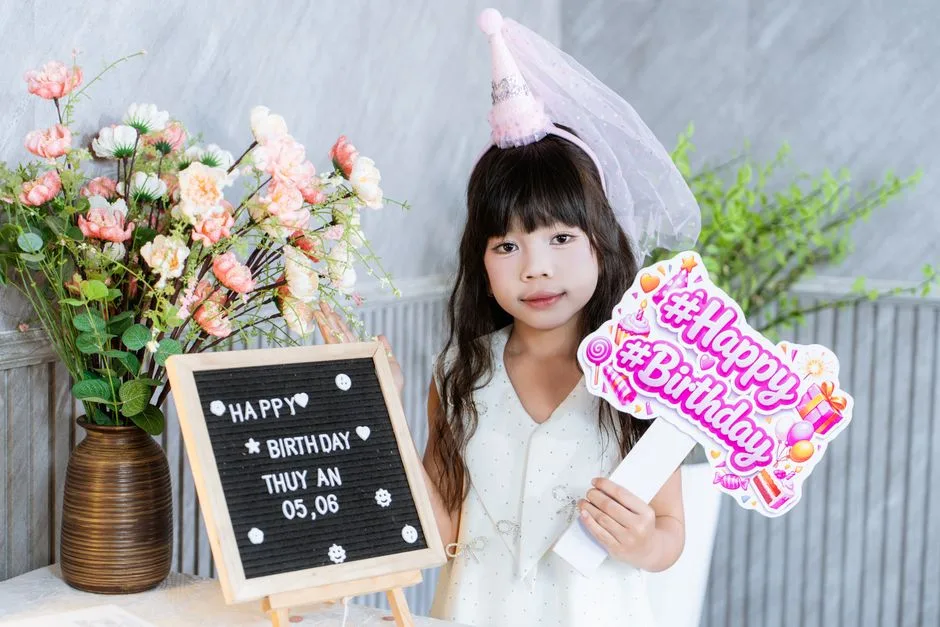 Young girl celebrating birthday indoors with flowers and sign.