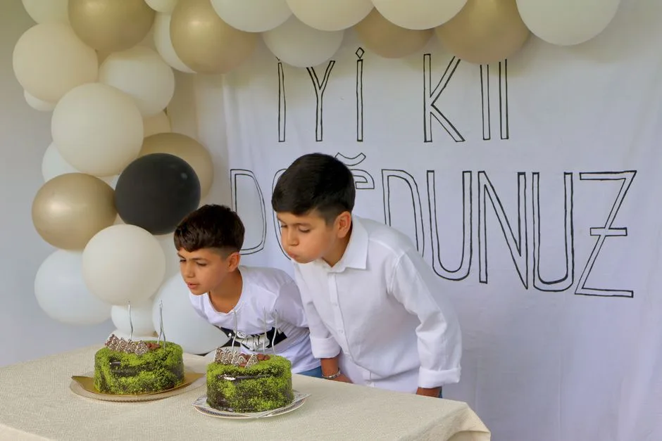 Boys blowing out candles on birthday cakes under decorated arch with balloons.