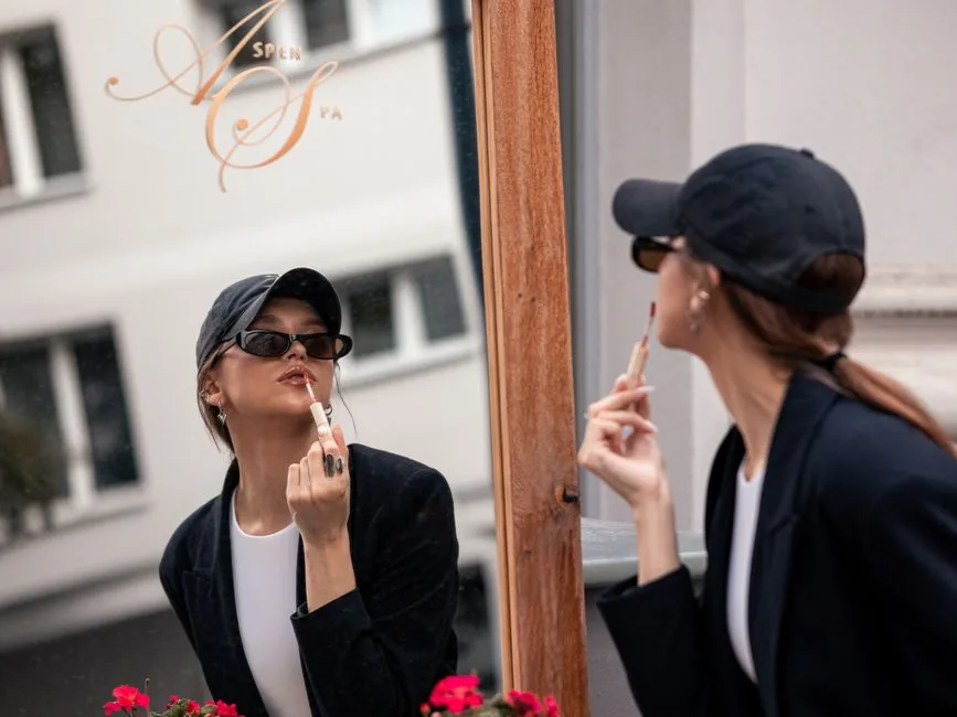 Stylish woman in cap applying lip gloss while gazing into mirror outdoors.