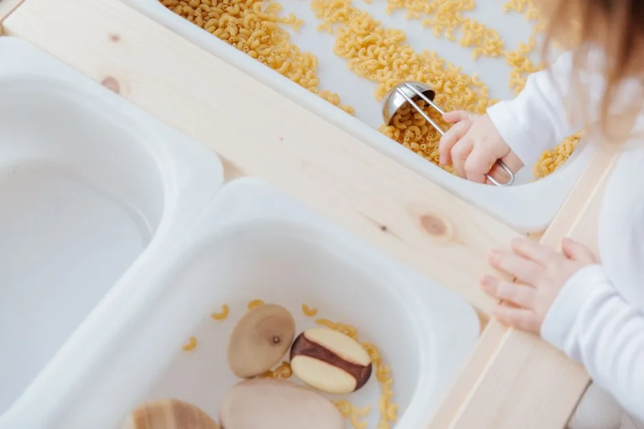 From above view of anonymous little kid taking raw pasta with metal spoon from white plastic container while playing at home