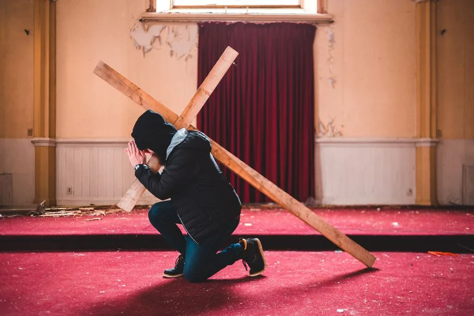 A person kneeling in prayer, carrying a wooden cross inside an abandoned chapel with red carpet.