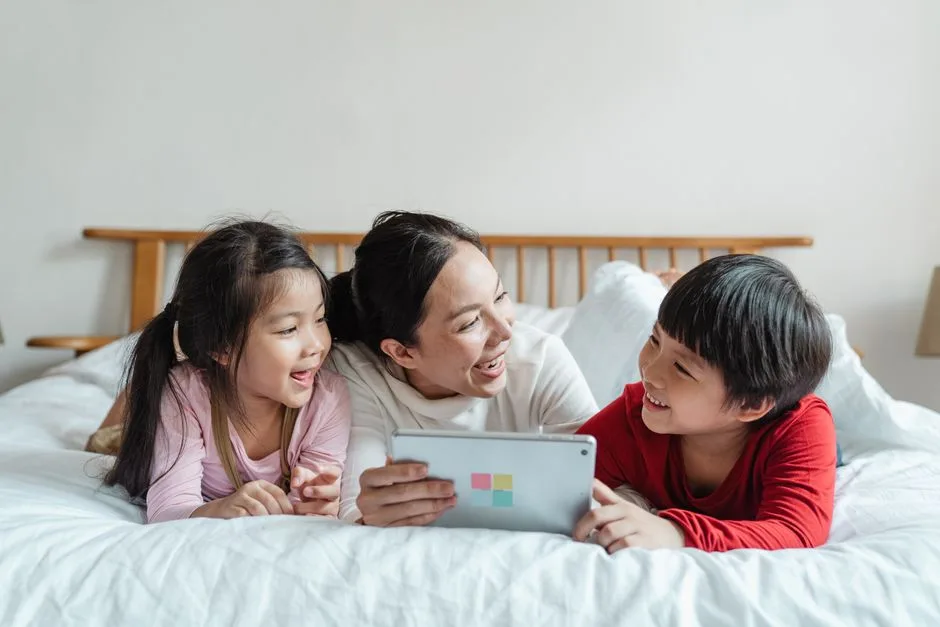 Full body happy Asian female with smiling little children lying on belly in casual clothes on soft bed while using tablet in light apartment
