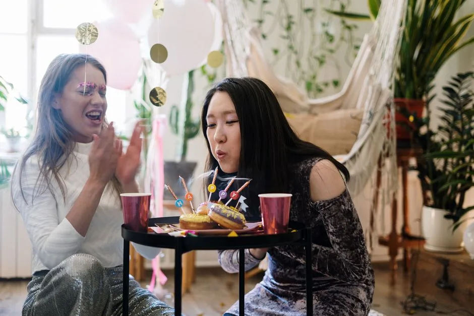 Asian female blowing candles with friend at a vibrant indoor birthday party featuring sweets and decorations.