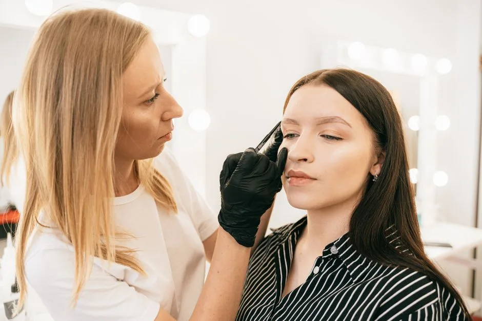 A skilled makeup artist applies eyebrow makeup on a client in a light-filled studio.