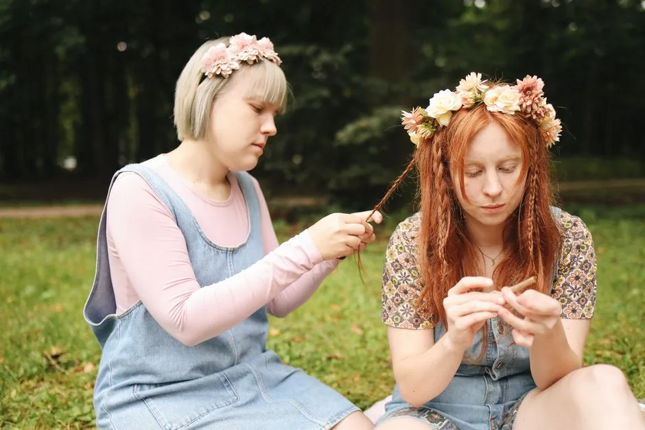 Two young women sitting outdoors, braiding hair and wearing flower crowns.