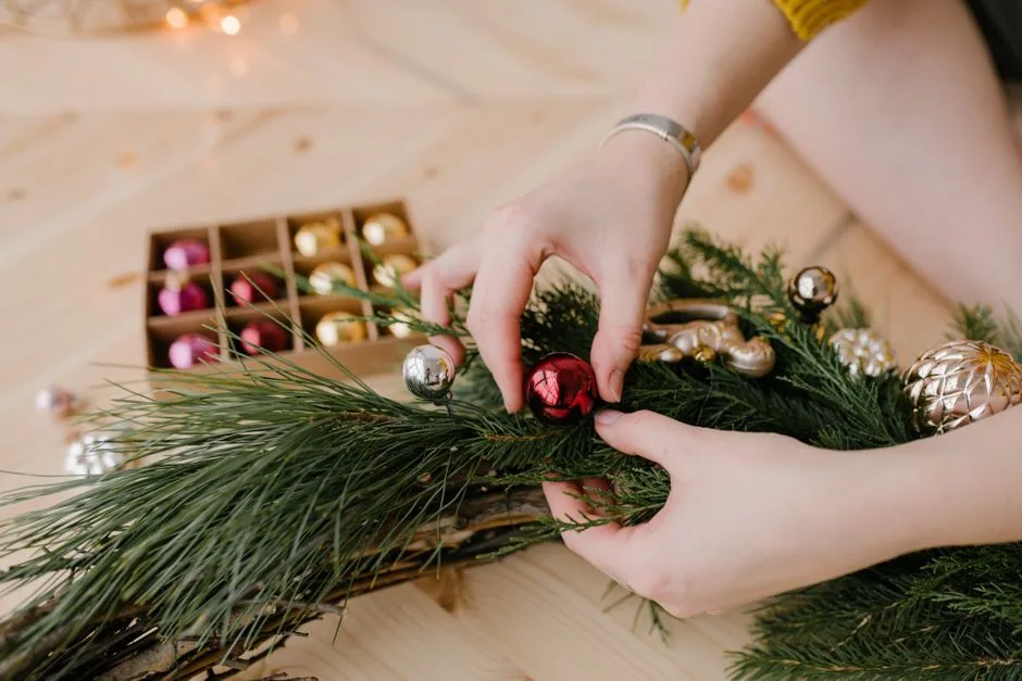 Hands crafting a Christmas wreath with colorful ornaments on a wooden surface.