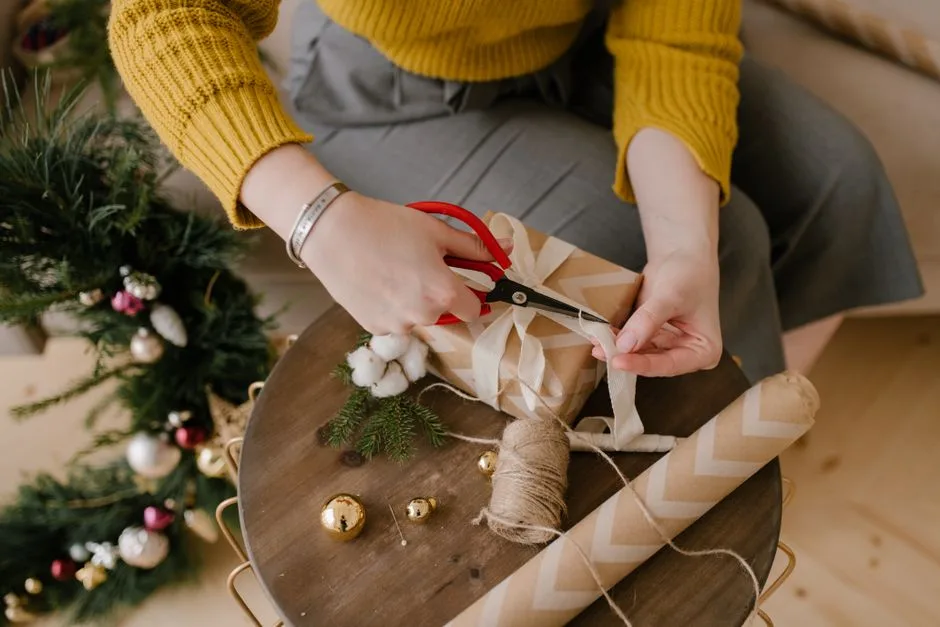 An adult wrapping a Christmas gift with ribbons and decorations at home.