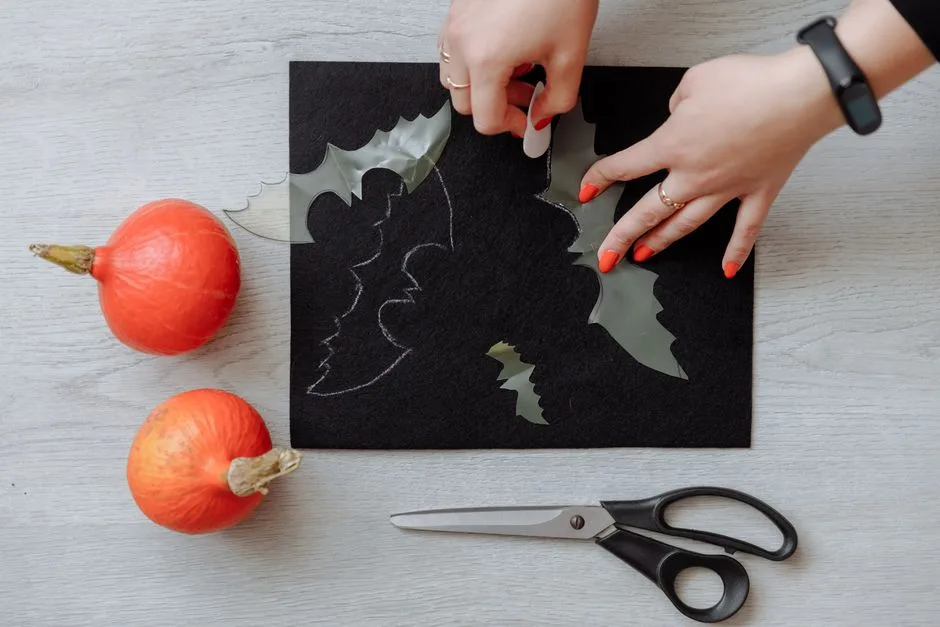 Hands crafting Halloween bat decorations on black paper with scissors and pumpkins.