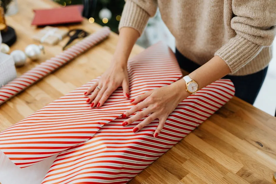 A woman wraps a Christmas gift with striped paper on a wooden table indoors, festive and cozy vibe.