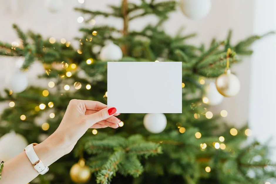 Hand holding a blank card with a decorated Christmas tree in the background, perfect for holiday greetings.