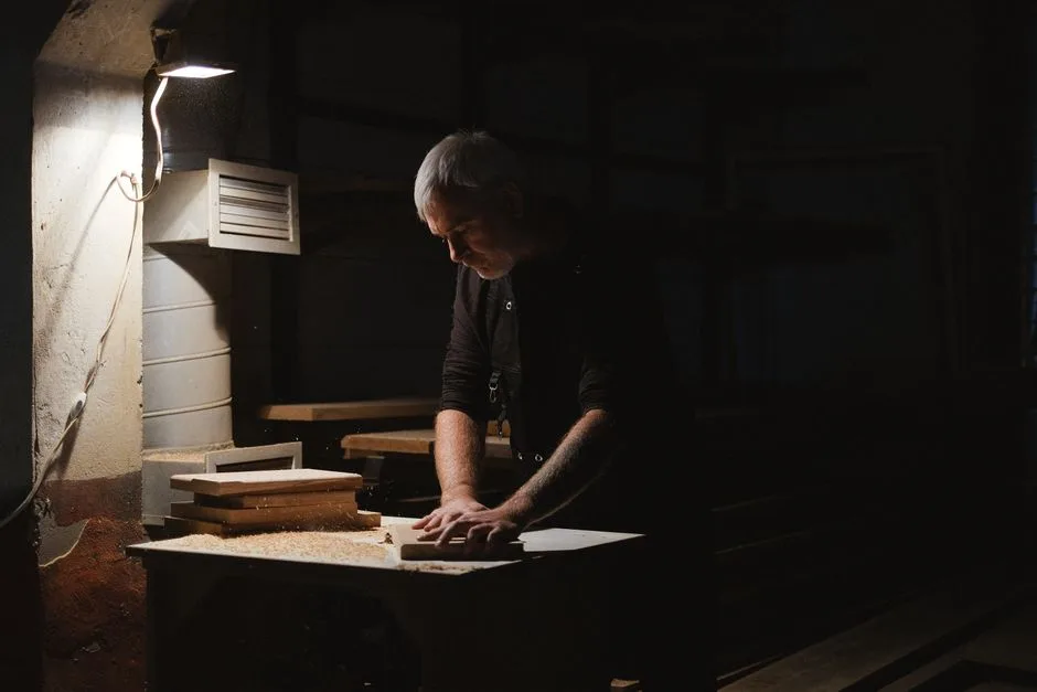 Side view of attentive male carpenter standing at router table while shaping wooden detail in dark workshop with glowing lamp
