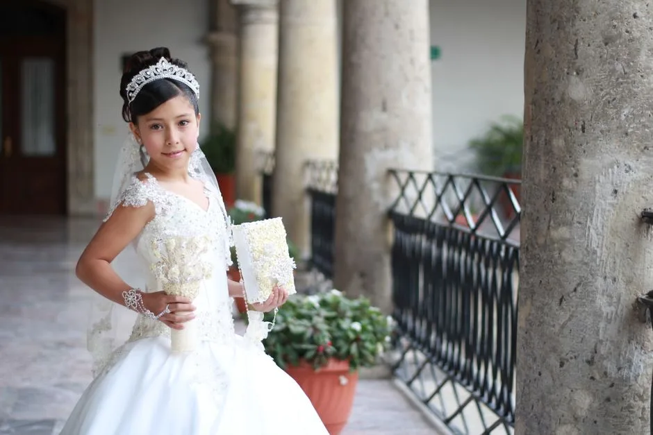 A young girl poses in a traditional white dress for her first communion ceremony, holding a bouquet.