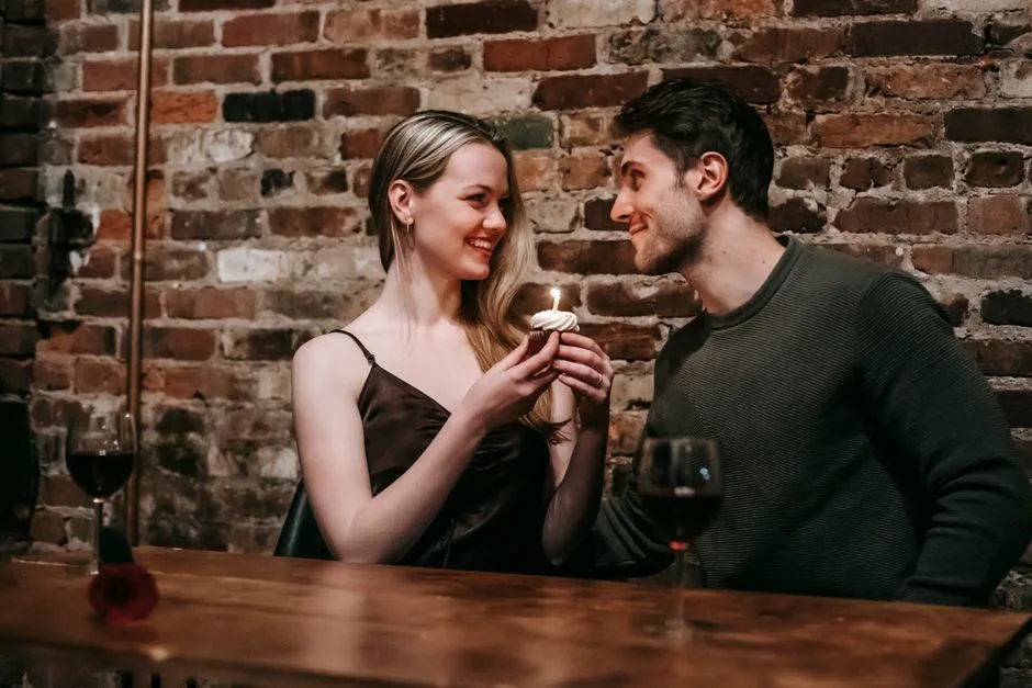 Side view of loving couple sitting near brick wall at wooden table with cupcake with burning candle looking at each other while celebrating anniversary in cafe