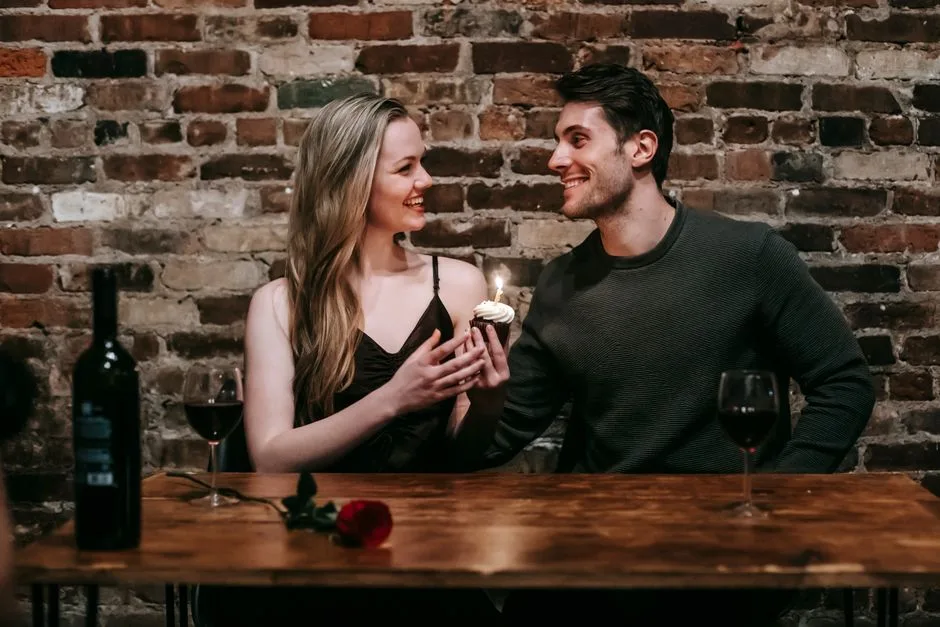 Side view of content couple with burning candle sitting near brick wall at wooden table with wine and rose in restaurant and looking at each other while spending romantic date
