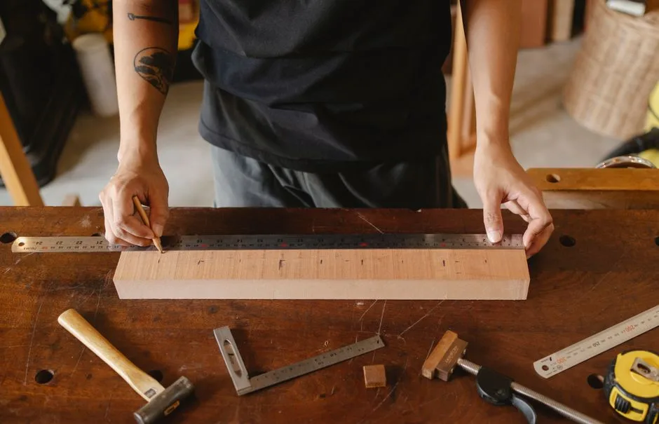 Crop anonymous tattooed male working with ruler and pencil while measuring timber plank on table with hammer and tools