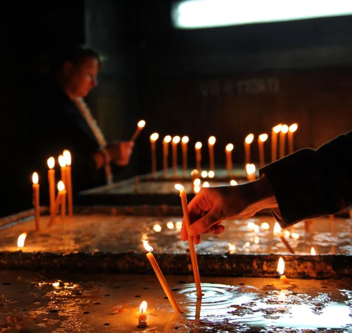 Crop unrecognizable person holding flaming candle and putting on stand while praying in cathedral