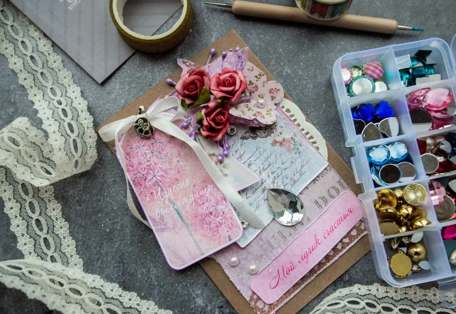 Top view of handicraft greeting card decorated with artificial flowers and fancy stones placed on table near box of colorful beads and buttons and tape lace